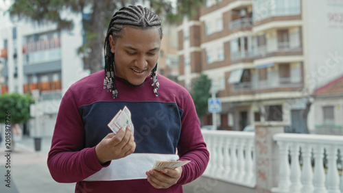 Canvastavla Young man counting pesos banknotes with both hands, smiling and looking down beside a white balustrade and apartment buildings on a city street; calm confidence
