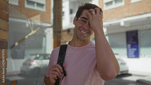 Fotografie Man smiles, runs hand through hair and holds backpack strap on a sunlit urban street with parked cars; casual ease relaxed