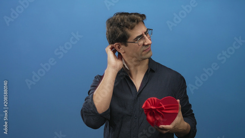 Obraz na płótnie Man with glasses touching cheek while holding red heart gift box in studio against blue backdrop; longing romance