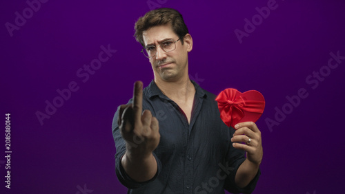 Fotografie Man in dark shirt holds a red satin heart gift with bow while showing middle finger toward camera in purple studio; defiance rejection