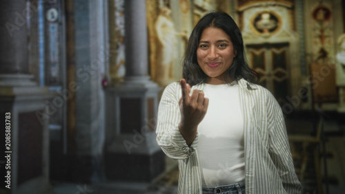 Tableau sur toile Woman showing middle finger gesture with bare hand and slight smile while wearing white shirt and striped overshirt in ornate building interior; defiance