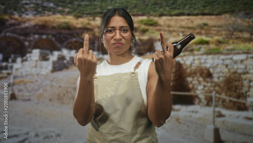 Quadro su tela Young woman in apron holds beer bottle and shows middle finger at an ancient ruins building; defiance protest