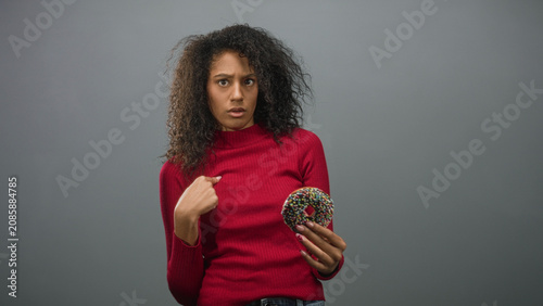 Konstfotografi Woman with curly hair wearing red sweater holding chocolate doughnut with sprinkles and pointing finger to chest in studio; temptation disbelief