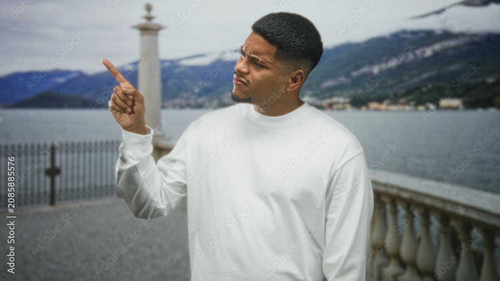 Fototapeta premium Hispanic man with beard in white sweatshirt points index finger upward on a lakeside street by a stone balustrade and iron railing with mountains across the lake; confidence.