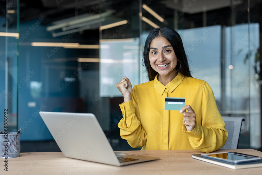 Fototapeta premium Happy young Indian woman sitting at office desk, holding credit financial card in hand and showing success gesture