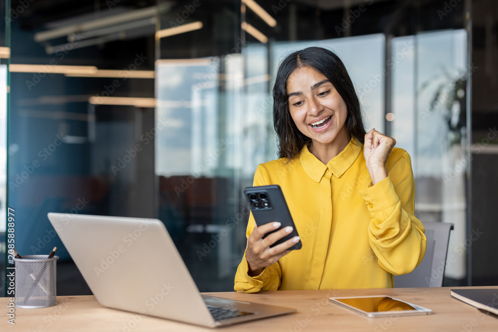 Fototapeta premium Happy woman celebrating success on the phone, sitting at the desk in the office and showing a victory sign with her hand