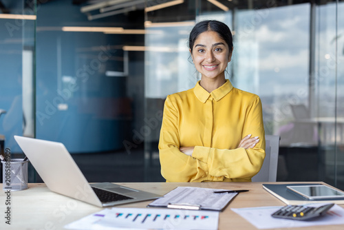Portrait of a young Indian businesswoman sitting in the office at a desk, with documents and a laptop, arms crossed on her chest and smiling at the camera
