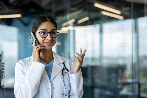 Smiling young Indian doctor standing in clinic office and talking on phone