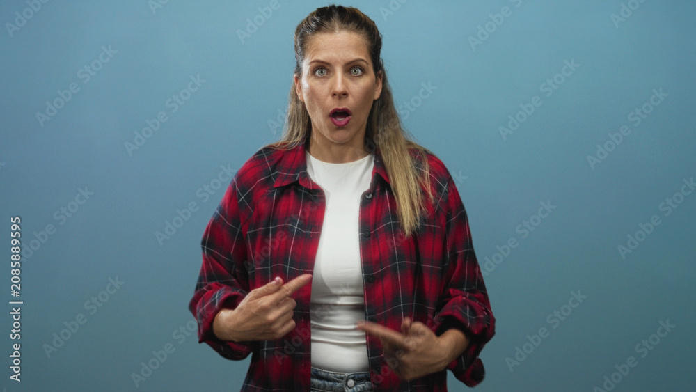 Obraz premium Woman in red plaid shirt pointing at herself with both thumbs and wide open mouth in studio; surprise disbelief.