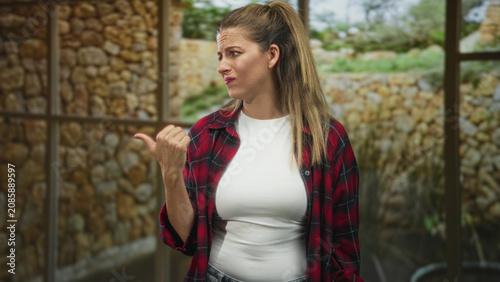 Fotografi Woman pointing thumb over shoulder inside building, wearing red plaid shirt and white tee; annoyance refusal