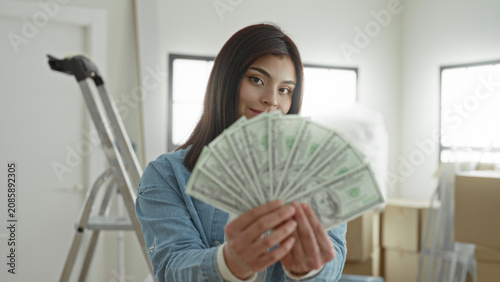 Εκτύπωση καμβά Young hispanic woman holds fan of dollars with hands, smiling amid packed moving boxes and ladder in sunlit building; moving day joy