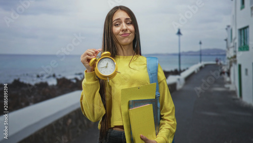 Fotografie Teenage student holds a yellow alarm clock and school folders on a street by the sea, lifting the clock with a smirk and raised wrist gesture; time pressure playful