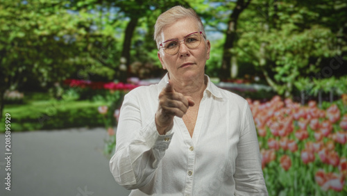 Tableau sur toile Woman points finger toward camera in forest setting beside tulip beds wearing glasses and white shirt; authority