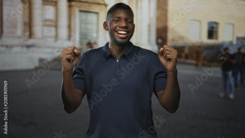 Photography Man smiling with clenched fists raised on a street in front of a stone building, eyes closed and elbows bent in a celebratory pose; joy
