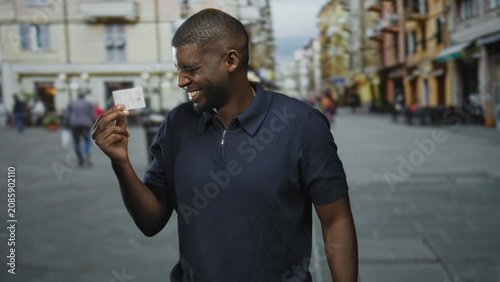 Fotografie Man holds creditcard between fingers on a street lined with buildings and cafe terraces while smiling at the card; confidence trust success
