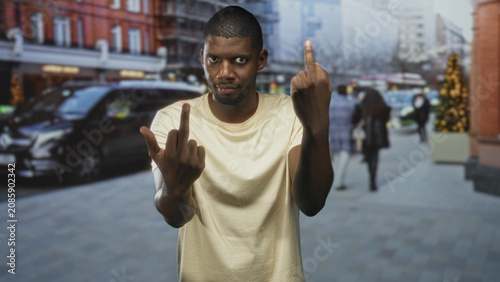 Obraz na płótnie Man showing two raised middle fingers toward camera while standing on a busy urban shopping street with parked car and decorated holiday tree; defiance provocation