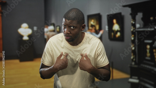 Photos Man pointing thumbs to chest with questioning frown amid framed paintings in museum building, defensive posture and raised eyebrows; confusion doubt