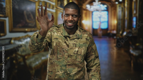 Schilderij op canvas Soldier waves hand and smiles inside an ornate building hall wearing camouflage uniform and raised palm gesture; pride duty welcome