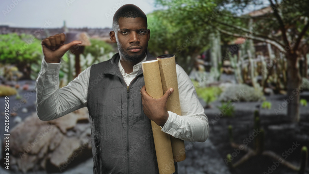 Fototapeta premium Man holding rolled blueprints and tubes, thumb to chest, wearing vest, cactus backdrop in studio; confidence planning design.