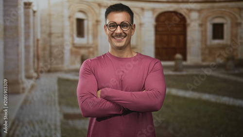Tableau sur toile Young hispanic man with moustache and glasses, arms crossed and smiling in building courtyard wearing pink shirt; confidence ease