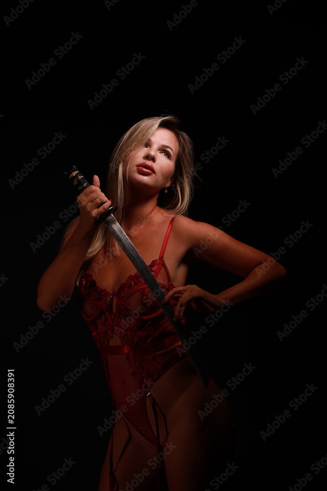 Obraz premium Low key boudoir portrait of a beautiful young woman with light brown hair in sheer red lingerie holding an authentic antique wakizashi sword