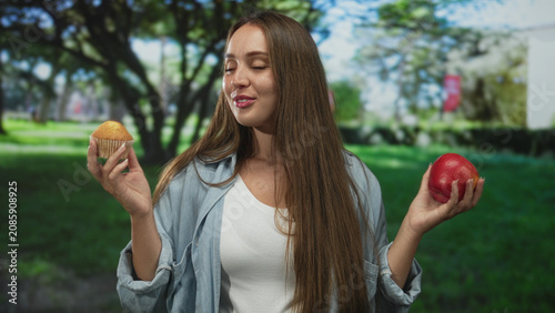 Photos Woman holding muffin and apple in a forest setting, eyes closed and savoring taste with both hands raised; temptation choice
