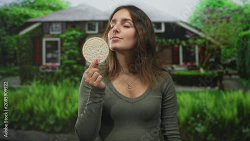 Fotografie Woman holding ricecake between fingers at house entrance building, smiling while presenting the round snack to camera; healthy snack contentment