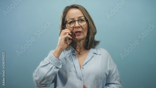 Fotografía Hispanic senior woman holding smartphone, fingers tapping screen while bringing phone to ear in studio; concern and focus