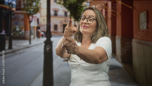 Quadro su tela Grey haired woman points finger like a pretend gun on a city street in front of a building; playful gesture
