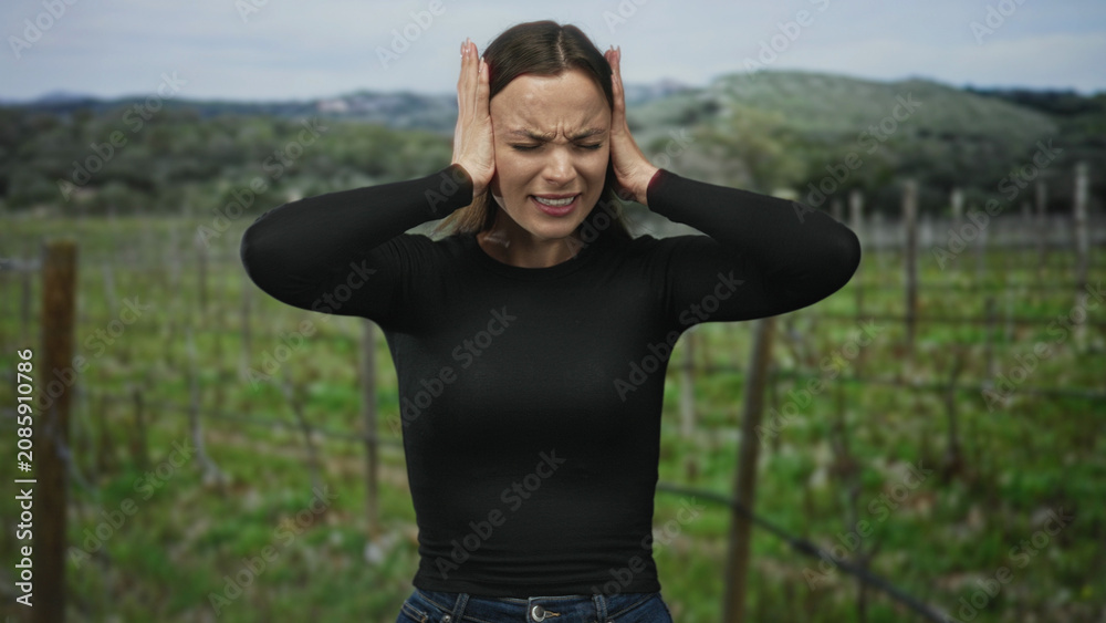 Obraz premium Woman in black top and jeans pressing hands to ears, eyes closed and mouth open in forest vineyard; emotional distress.