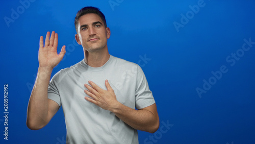 Fotografie Young hispanic man with hand on chest and raised hand, wearing light gray tshirt, direct gaze in blue studio; gratitude pledge
