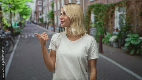 Fotografie Young blonde woman points thumb to street while smiling, visible hand and forearm, wearing plain white t shirt and casual summer look; friendly invitation