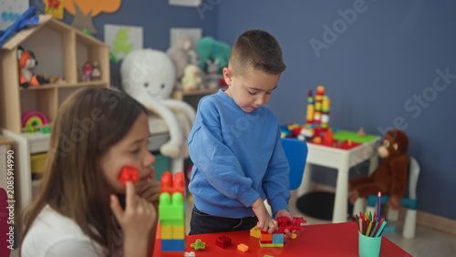 Photography Boy stacking colorful plastic blocks with hands on a red table in a playroom inside a building while girl watches and crayons sit in a cup; quiet concentration