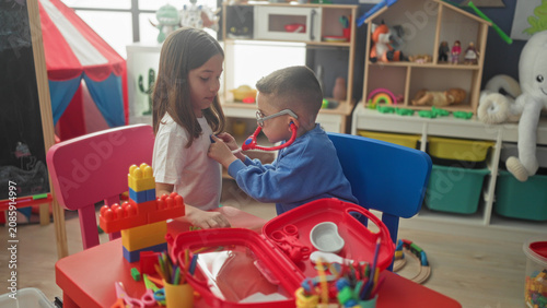 Fotografie Girl and boy roleplay doctor with red stethoscope, boy listens to girl's chest and adjusts earpiece at a playroom; playful care