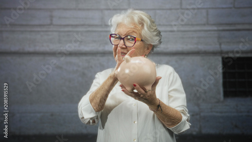 Foto Elderly woman holds piggy bank and gives thumbs up in front of building; savings independence optimism
