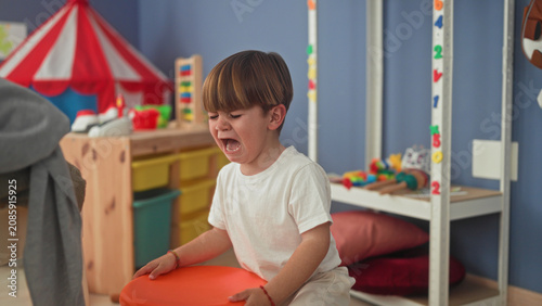 фотографія Boy crying with open mouth in a colorful playroom near a tent and toy shelf, reaching toward scattered toys and a pillow; tantrum sadness