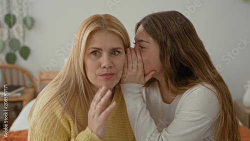 Photography Blonde mother listens as her daughter whispers into her ear with hand covering mouth while seated on a bed in a bedroom; surprise intimacy trust