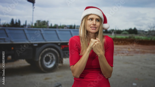 Foto Woman clasping hands near chest in santa hat beside flatbed truck on street with dirt lot and cloudy sky; hopeful plea