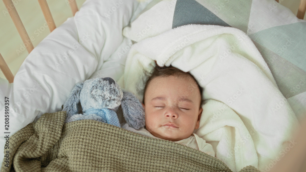 Naklejka premium Baby boy sleeping with closed eyes in a crib, face visible, swaddled under a green blanket beside a plush rabbit inside a building; serene rest.