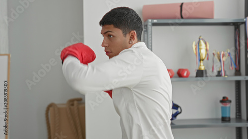 Φωτογραφία Man in white jacket throwing a jab with red boxing gloves in a studio, trophies and yoga mat visible; training focus determination