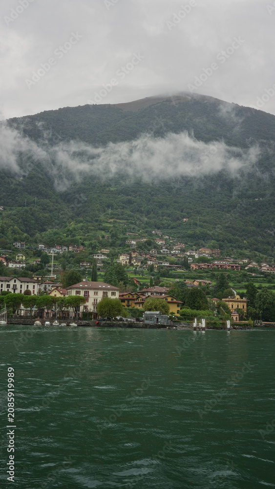Fototapeta premium Lakeside village with waterfront buildings, rippling emerald lake water and low clouds clinging to the mountain slopes; serenity.