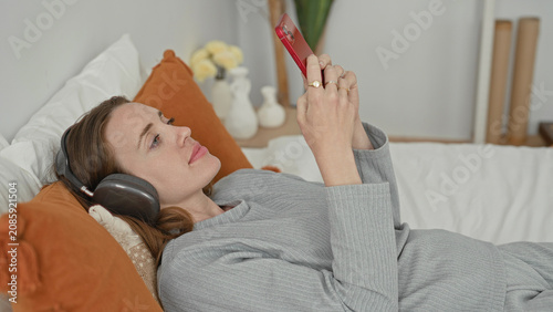 Bild auf Leinwand Woman lying on bed wearing headphones, holding smartphone aloft and tapping screen in building; calm connection