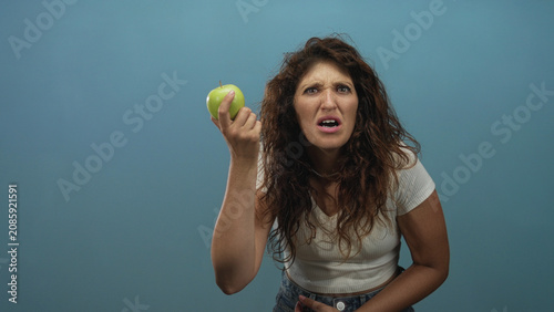 Photos Woman holding green apple in blue studio, hand raised and grimacing at the fruit while wearing white crop top; disgust confusion