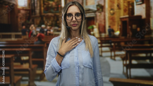 Obraz na płótnie Woman wearing headset and glasses with hand on chest among church pews and ornate altar interior, young hispanic blonde operator; surprise devotion