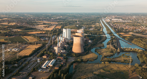 Industrial power plant with cooling towers surrounded by green fields and a canal under a clear blue sky, captured at sunset — symbol of modern energy and landscape harmony.