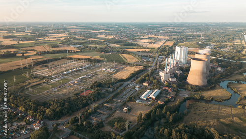 Industrial power plant with cooling towers surrounded by green fields and a canal under a clear blue sky, captured at sunset — symbol of modern energy and landscape harmony.
