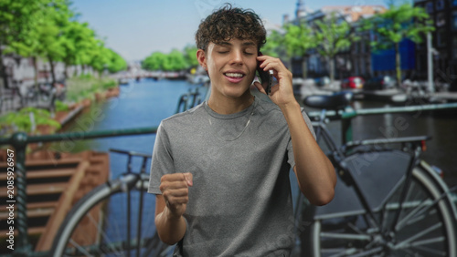 Tableau sur toile Teen boy on phone with hand to ear and a silver chain, smiling on a city street by a canal and bicycle; youthful joy