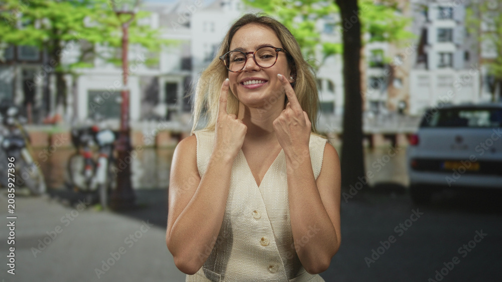 Fototapeta premium Woman smiling and pressing index fingers to cheeks on a street canal with parked bikes and car nearby; playful joy.