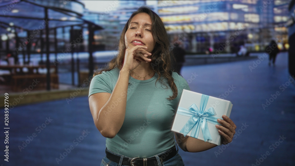 Fototapeta premium Young latina woman holding a gift box with hand on forehead on street in front of building, eyes closed showing discomfort for headache; disappointment fatigue headache.