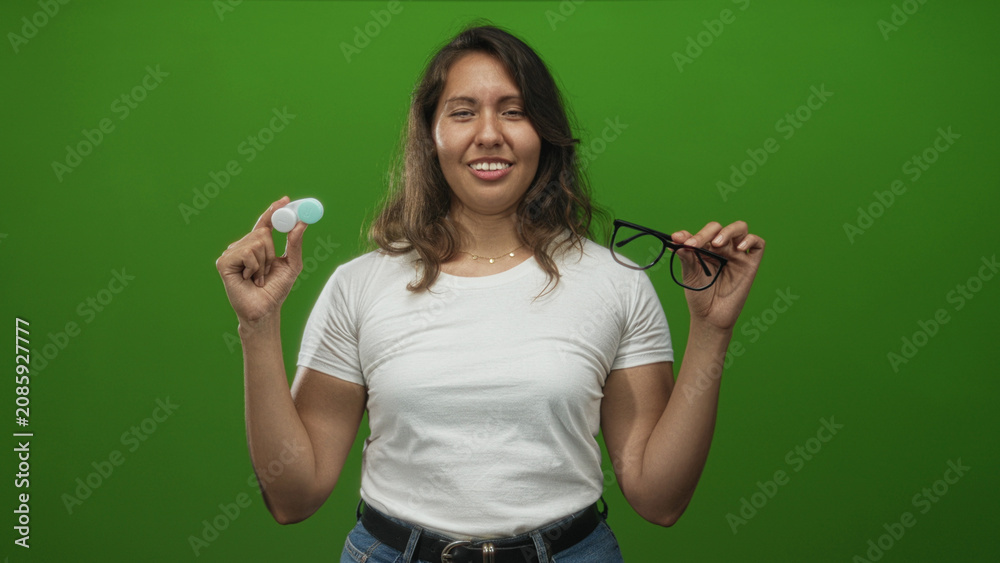 Fototapeta premium Woman holding a contact lens case and eyeglasses while squinting her eyes in a green studio setting showing indecision and comparison; playful choice.